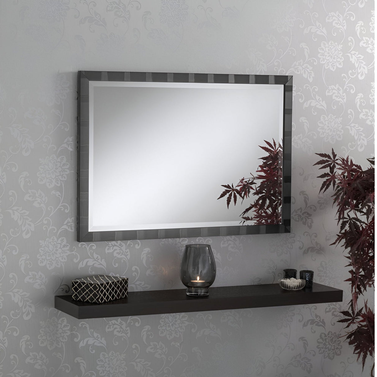 A modern interior features the Solvix Rectangle Wall Mirror - Black, stylishly installed above a floating shelf. The shelf displays decorative items such as a small potted plant and a candle in a dark holder. A subtle floral pattern embellishes the wallpaper, adding to the contemporary ambiance.