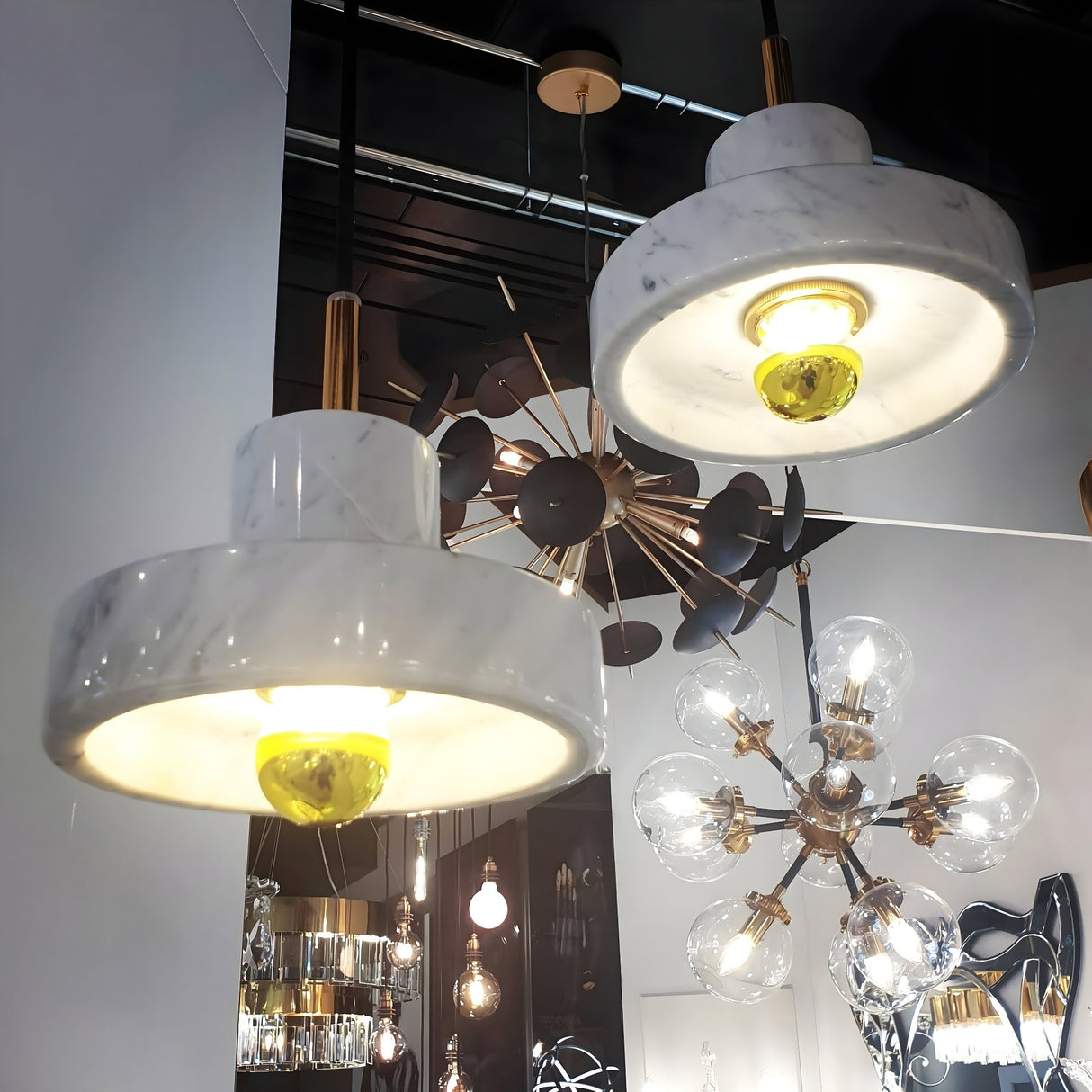 Two Marble Pendant Light - White & Gold fixtures with exposed bulbs hang before a reflective surface, surrounded by luxury lighting and contemporary gold-accented chandeliers in a stylish showroom.