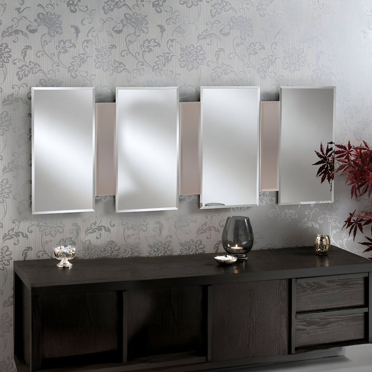 A modern interior showcases a dark wood sideboard with three drawers, above which a set of Miran Rectangle Wall Mirrors in bronze is arranged in a staggered pattern. Decorative items and a plant with red leaves adorn the sideboard, set against a floral wallpaper.