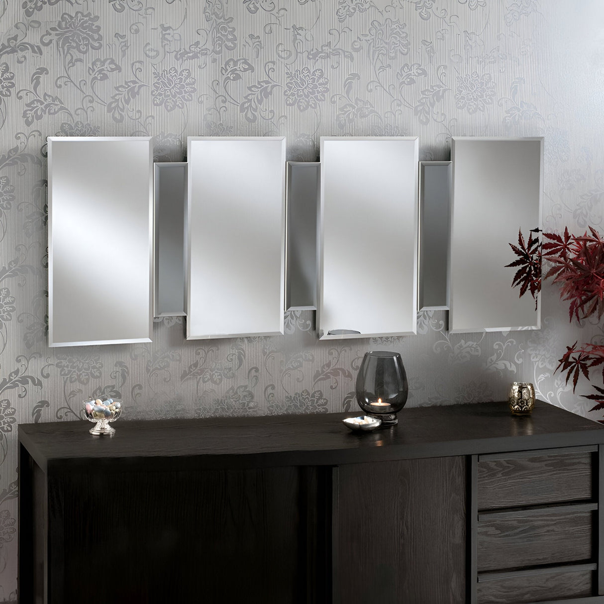 The Miran Rectangle Wall Mirror - Grey is mounted on a patterned wall. Below it, there's a dark wooden cabinet adorned with a glass candle holder and a small dish. To the right, a decorative red plant enhances the contemporary style's elegant ambiance.