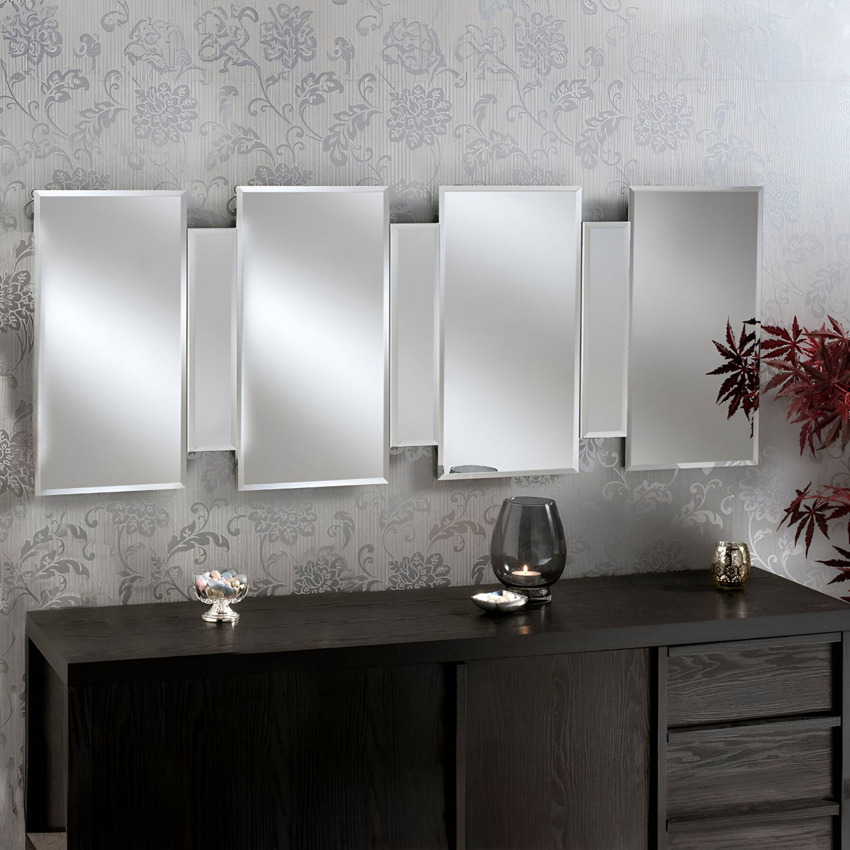 A contemporary Miran Rectangle Wall Mirror in silver, featuring varied rectangular panels, is mounted on a wall adorned with floral-patterned wallpaper. Below the mirror, a dark wood sideboard showcases a glass vase, a small plate, and decorative items. The setup is elegantly contrasted by a red-leafed plant.