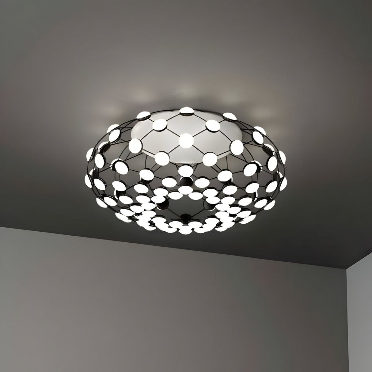 The Mesh LED Flush Ceiling Light - Black features a circular, geometric design with interconnected black metal and small round white bulbs, creating a modern and striking pattern on the ceiling.