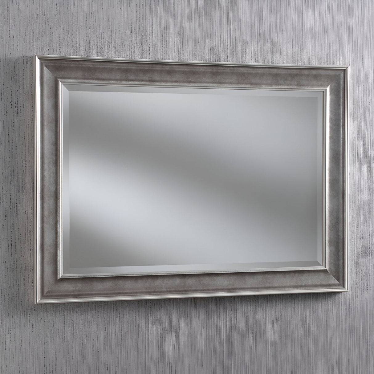 The Haloen Rectangle Wall Mirror - Silver is elegantly hung on a textured, light gray wall. This stunning piece of home decor reflects diffuse lighting, adding a modern touch to the setting with its wide silver frame.