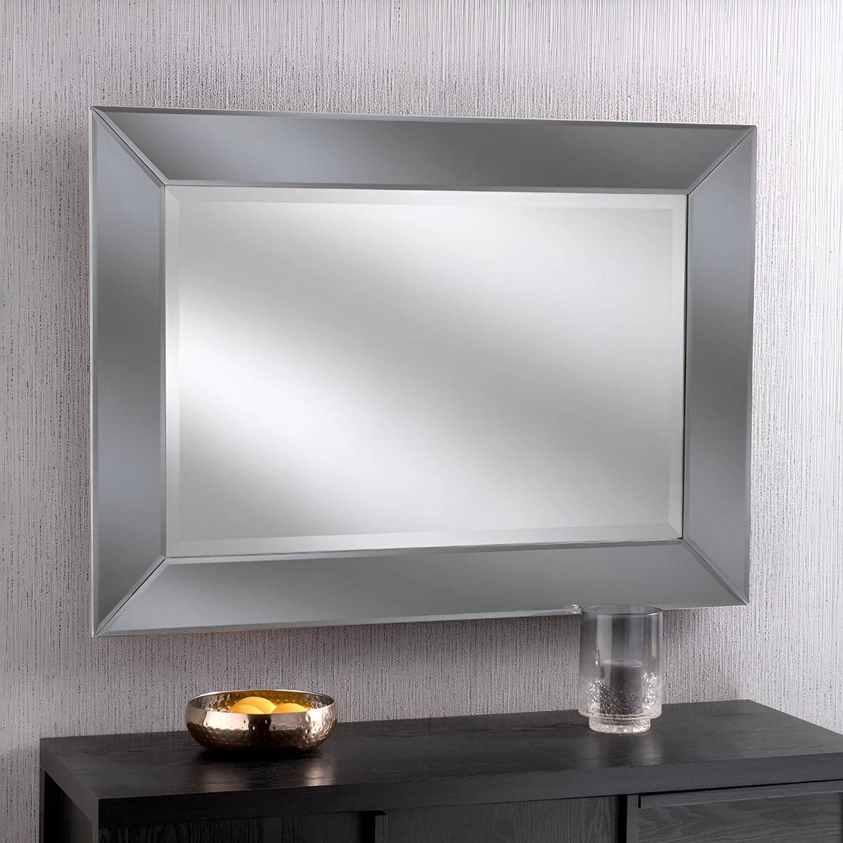 The Monaco Rectangle Wall Mirror in Grey (112x81cm) is mounted on a textured, light-colored wall, infusing the space with a modern flair. Positioned beneath it is a sleek black console table adorned with a circular metallic bowl and a clear glass tumbler, creating an air of contemporary elegance.