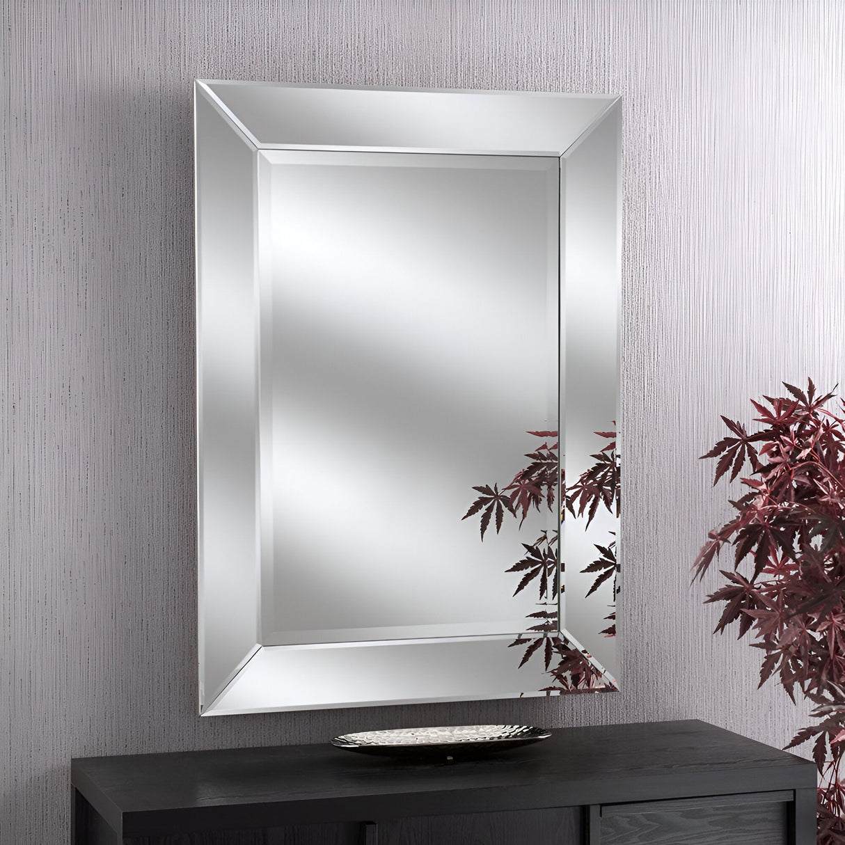 The Monaco Rectangle Wall Mirror Silver - 112x81cm, featuring a beveled edge, elegantly adorns a textured light gray wall. Below it is a dark wooden table with a decorative plate, and red leaves from a nearby plant are partially reflected in the mirror's silver frame.