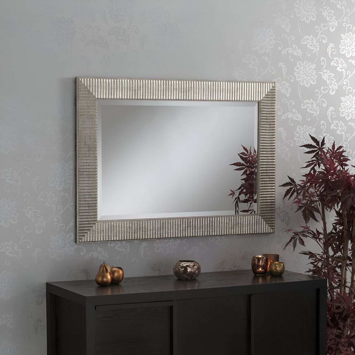 The Montana Rectangle Wall Mirror - Antique Silver, with its textured antique silver frame, hangs on a patterned wall, exuding vintage charm. Below is a black cabinet adorned with decorative items such as small metallic pumpkins and a silver vase. Beside the cabinet stands a plant with dark red leaves.