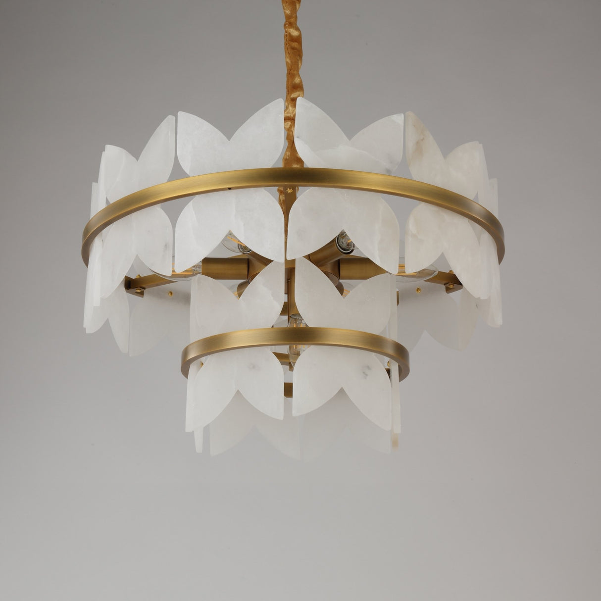 Niori Petraia two-tier chandelier with sculpted white alabaster petals on brushed brass rings, suspended from a fabric-wrapped chain against a grey background.