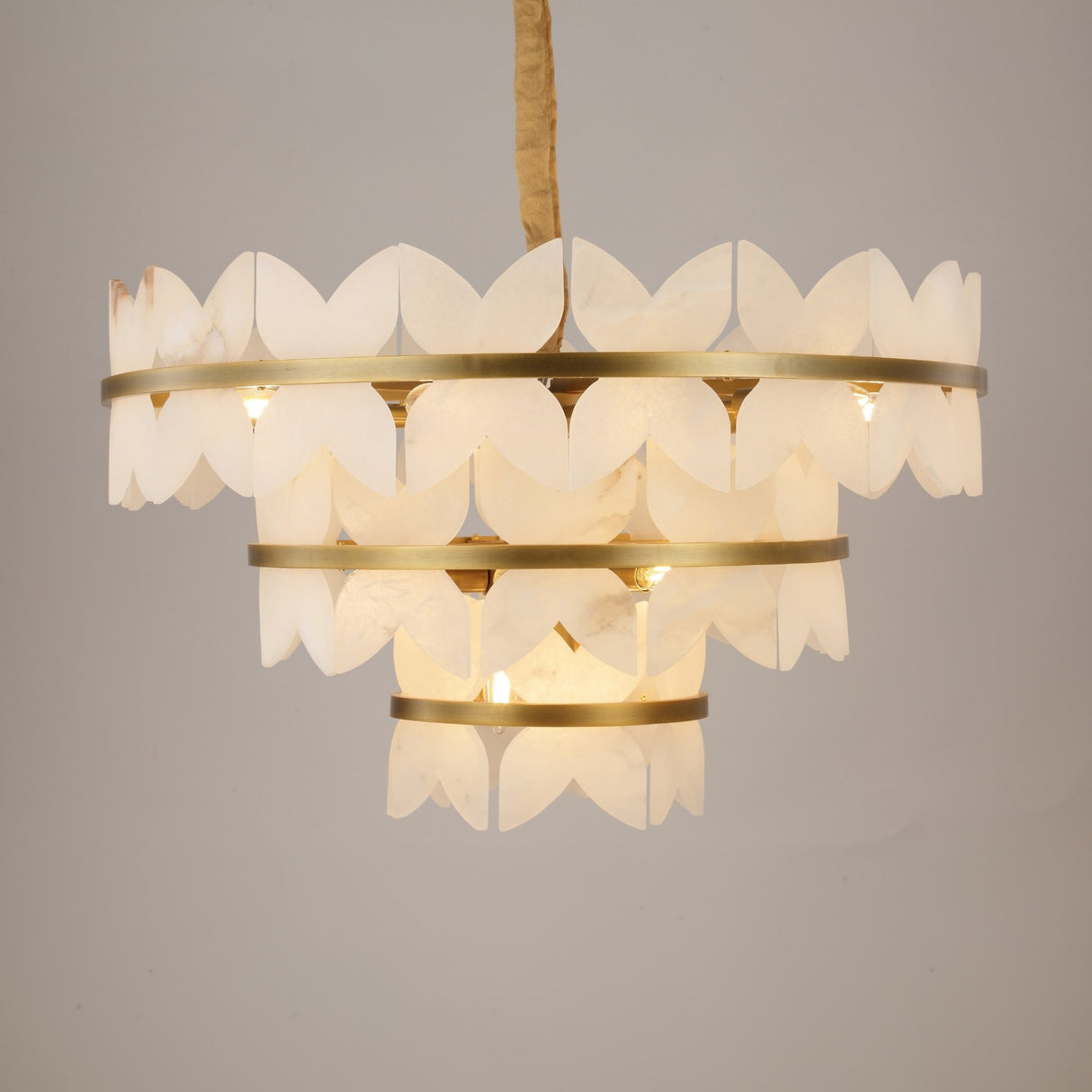 Niori Petraia three-tier chandelier with scalloped alabaster petals on brushed brass rings, suspended from a fabric-wrapped chain against a pale background.