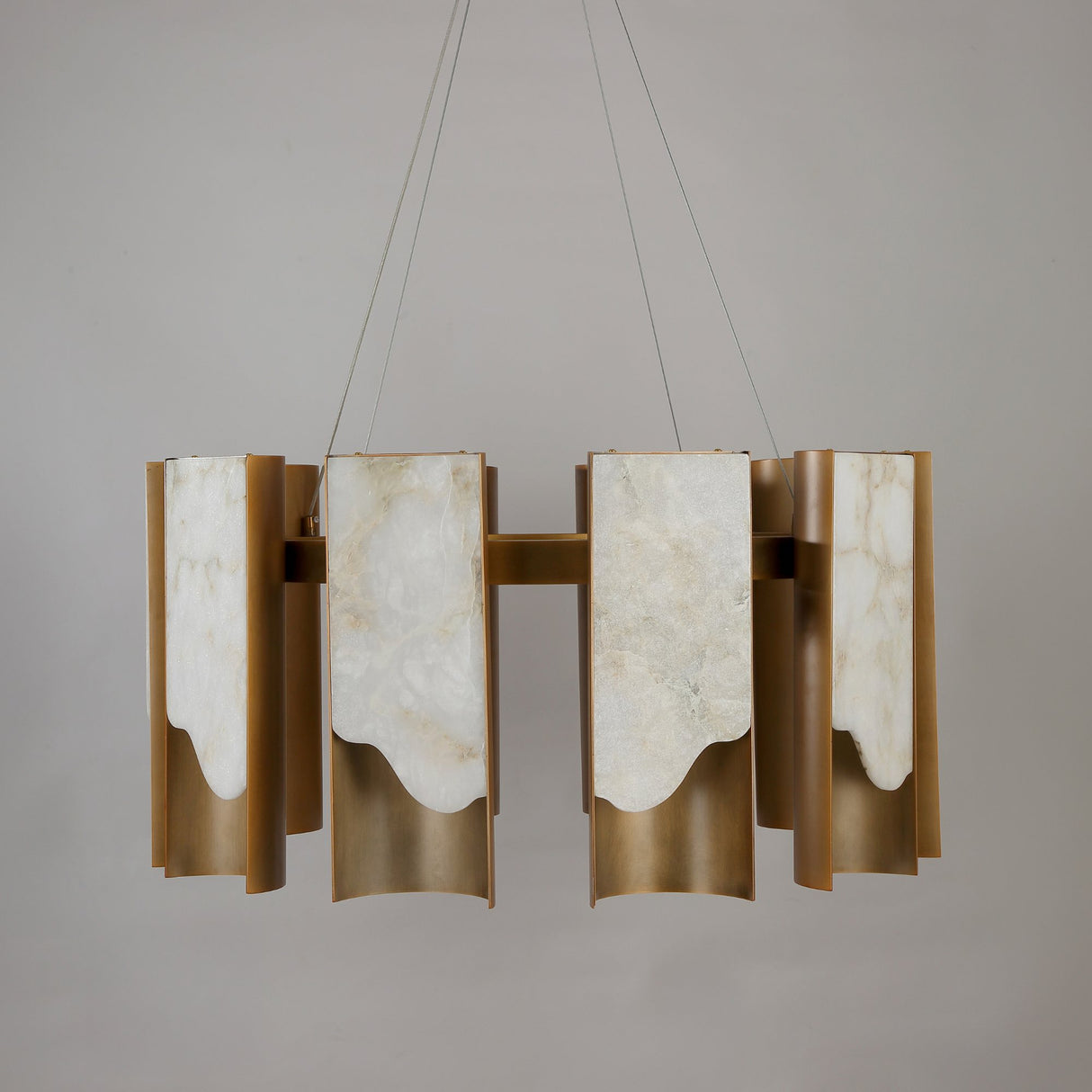 Niori Caelith LED large circular chandelier with stepped Spanish alabaster panels and brushed brass blades, suspended on steel cables against a grey background.