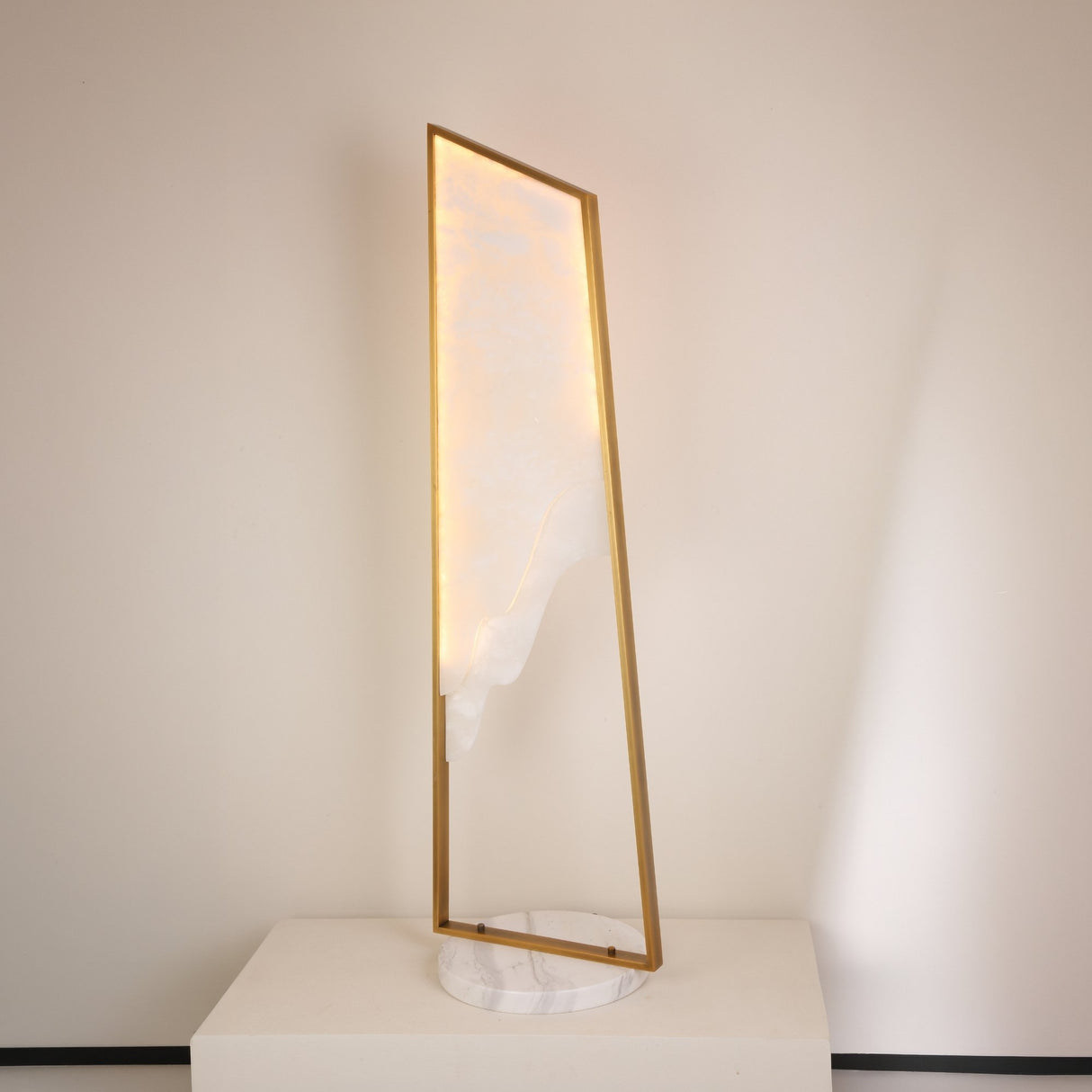 Niori Atria Small LED alabaster floor lamp with tapered illuminated panel in a brushed brass frame on a round white marble base, shown on a plinth.