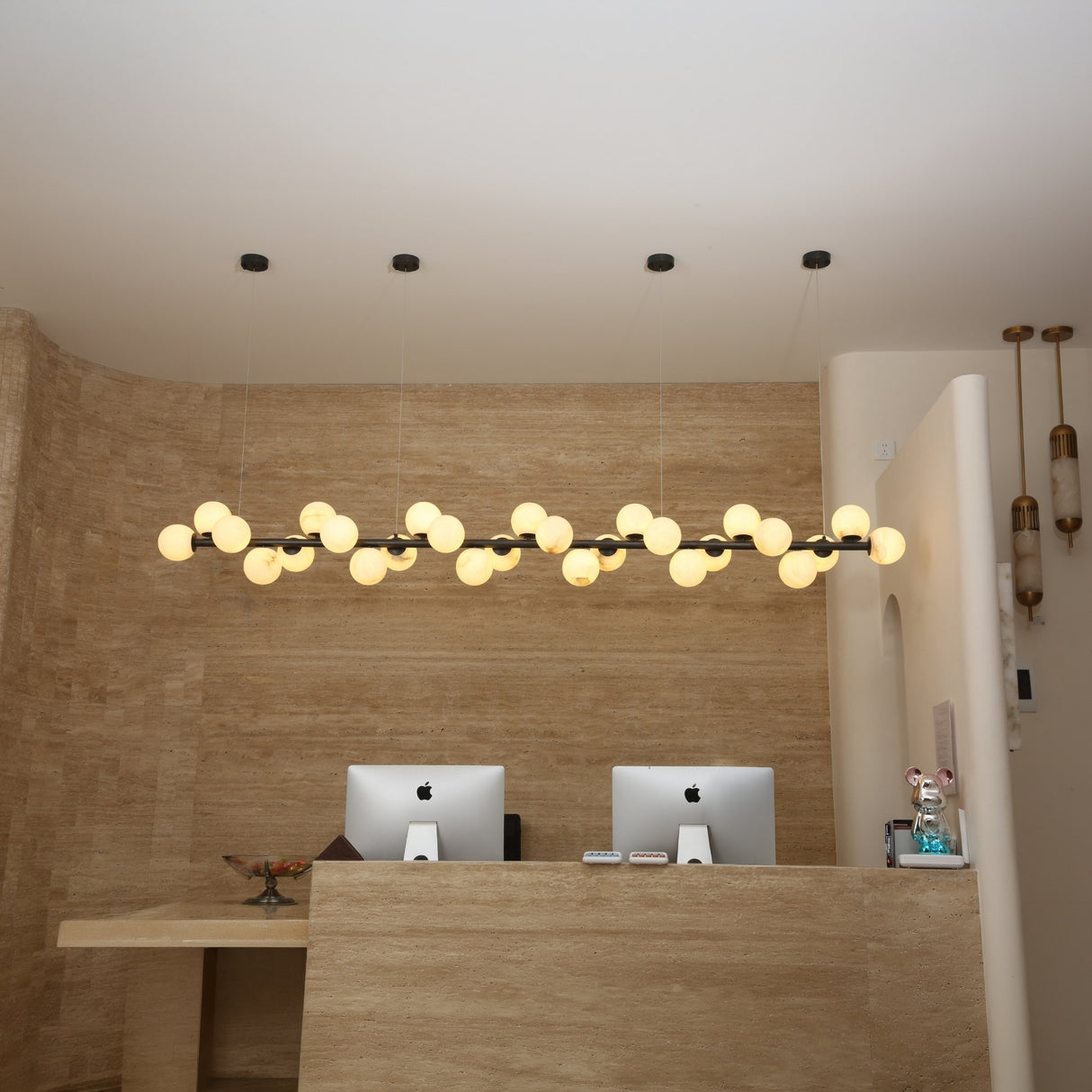 Niori Axis 27 Light Alabaster Linear Chandelier in matte black suspended above a travertine reception desk with iMacs in a contemporary interior.