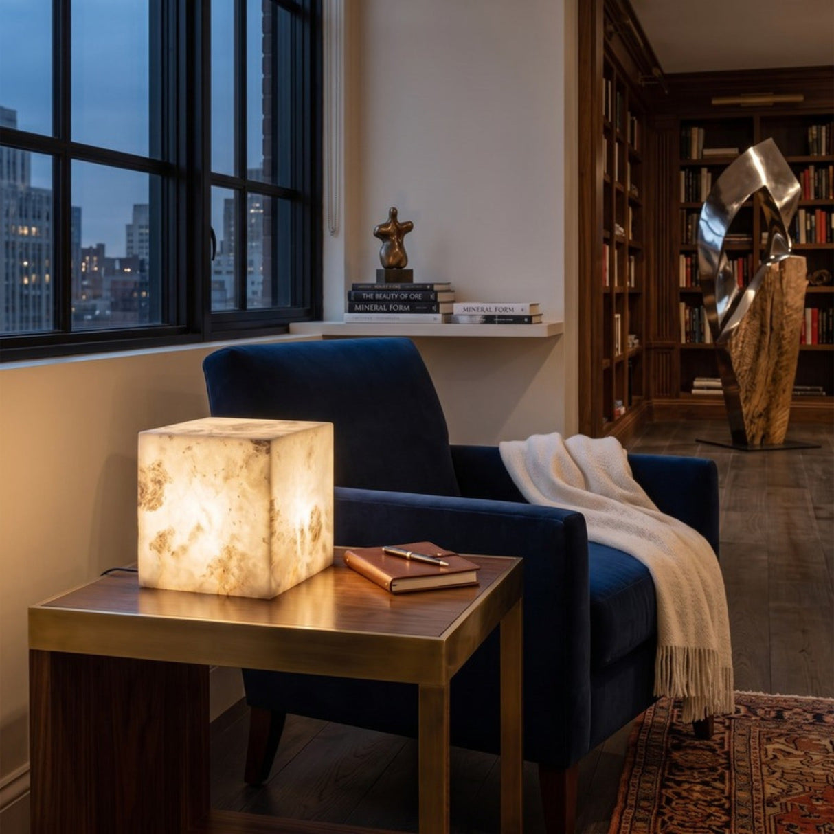 Blocchetto LED Cube Alabaster Table Lamp, 12cm glowing natural stone cube on a brass side table, shown in a living room beside a navy armchair.