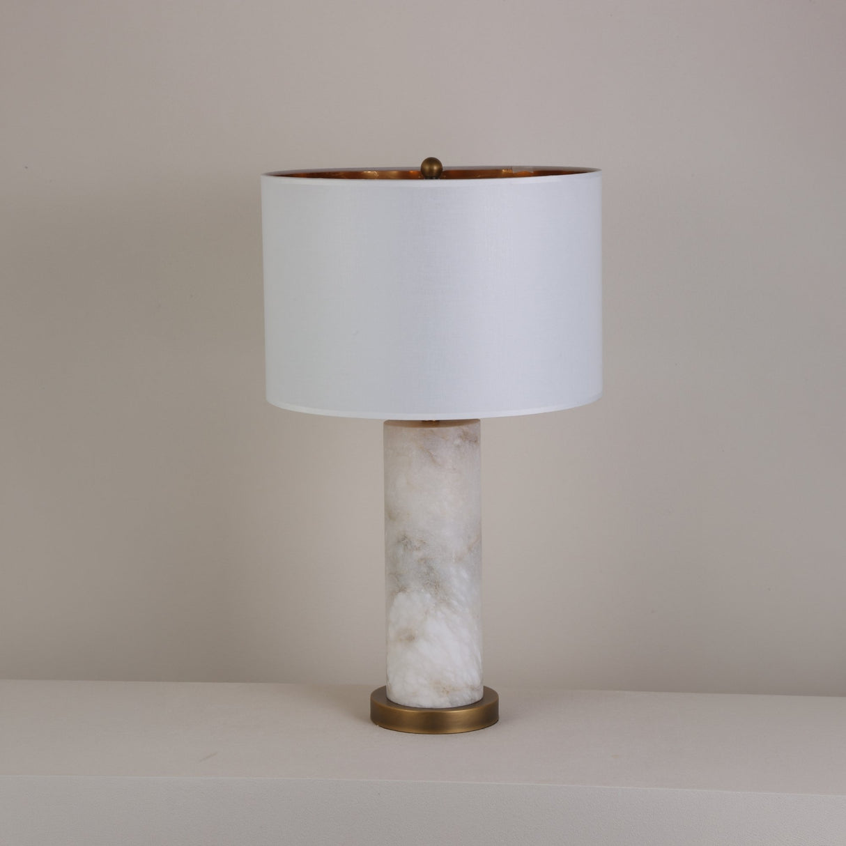 Calynor 1 Light Column Alabaster Table Lamp with brushed brass base and stem, topped with a white drum shade, shown against a plain neutral background.