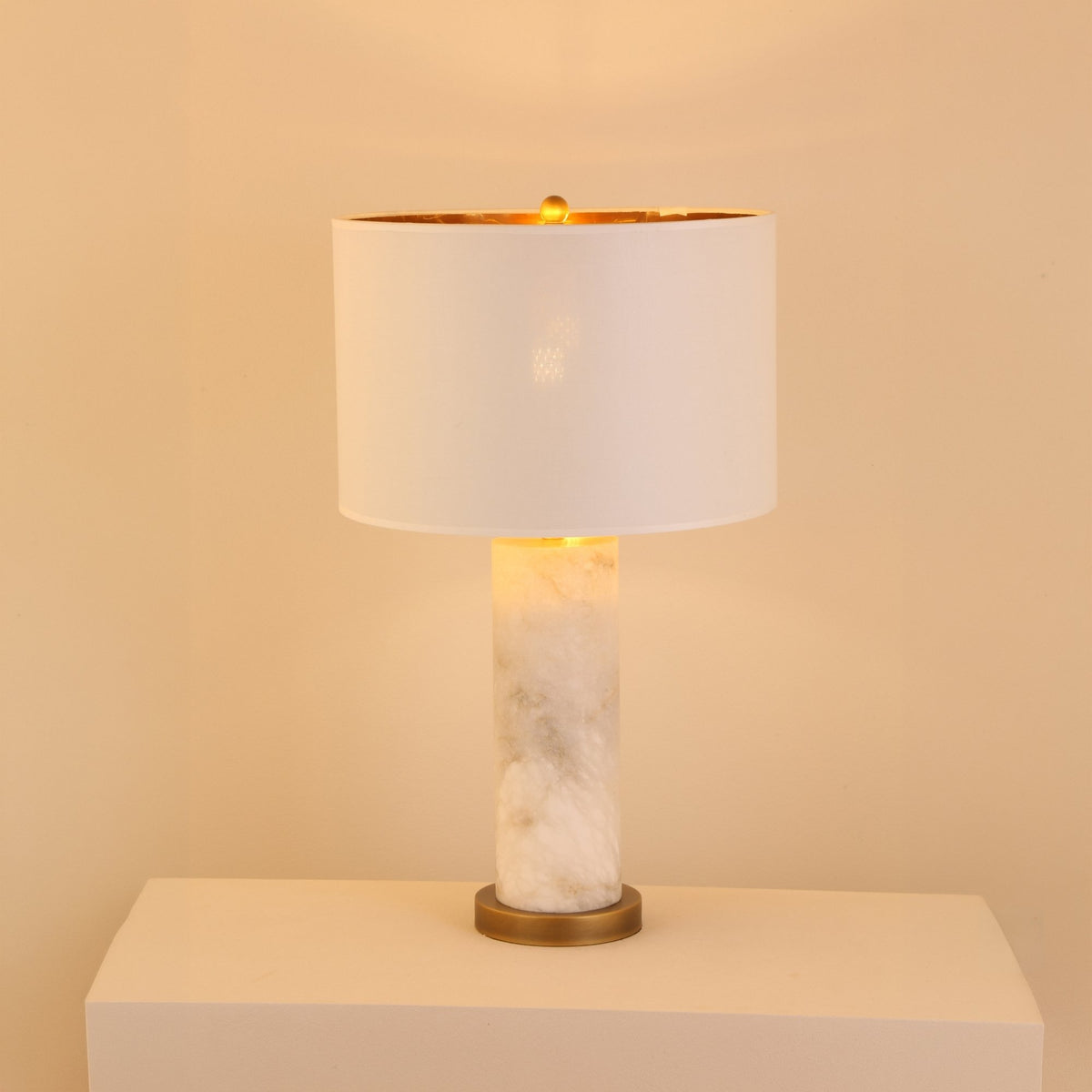 Calynor 1 Light Column Alabaster Table Lamp with white marbled stone base, brushed brass stem, and ivory drum shade, glowing softly against a plain light background.