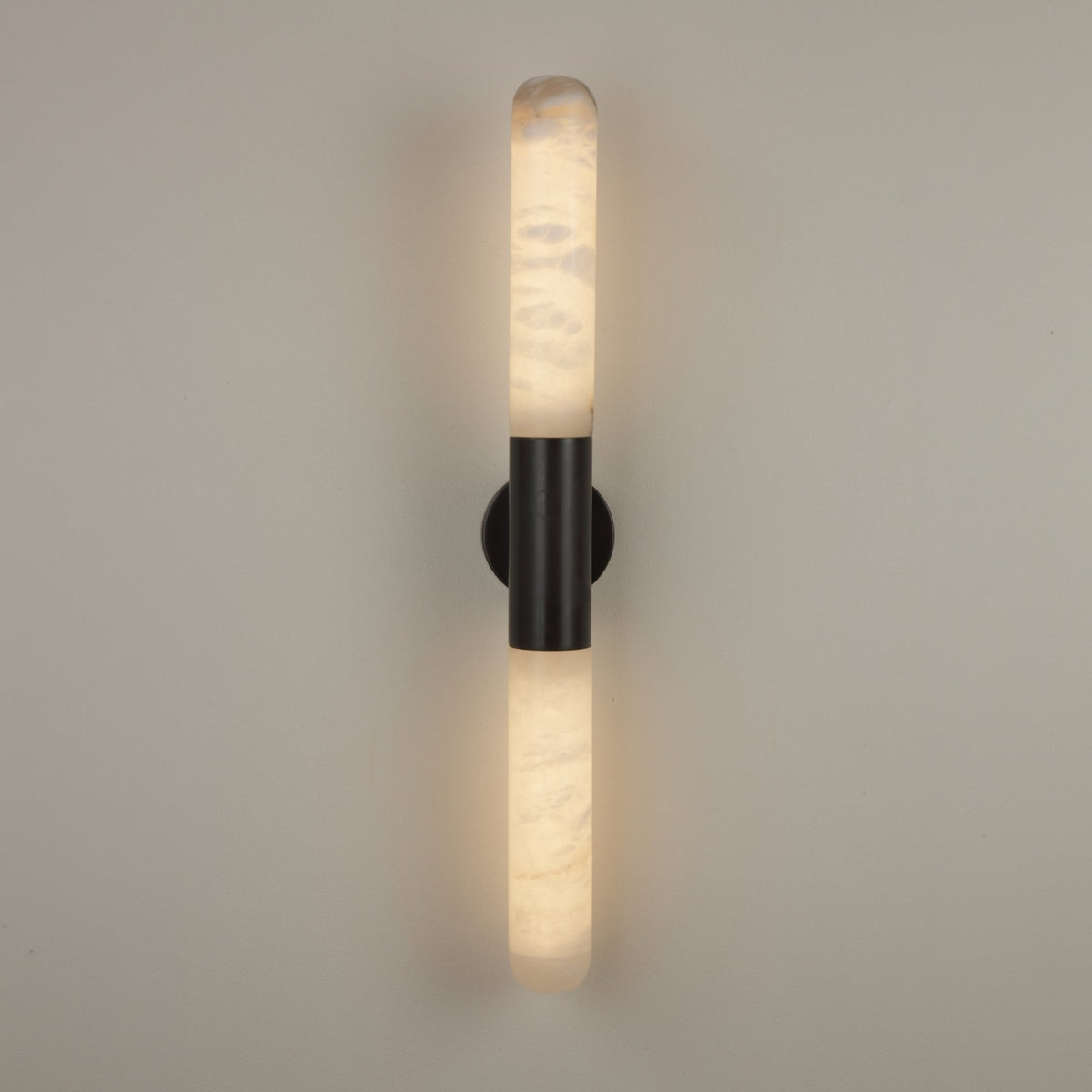 Averin LED Alabaster Wall Light in Matte Black, vertical fixture with two glowing alabaster columns joined by a black centre bracket, on a plain grey wall.