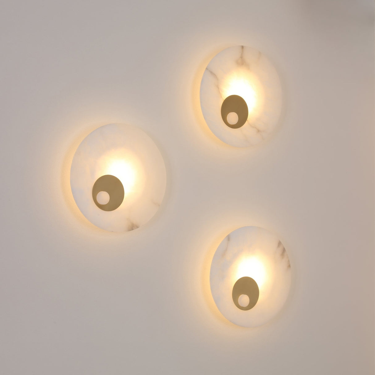 Three Selvana Small round alabaster wall lights with brushed brass centre bosses, mounted on a plain wall and glowing warm white when lit.