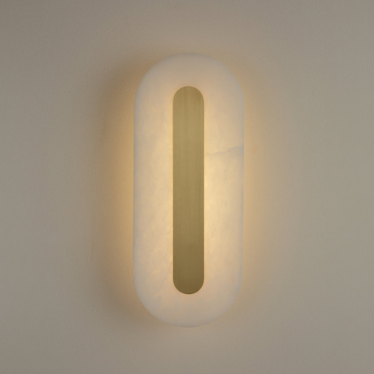 Averra medium alabaster wall light by Niori, illuminated pill-shaped panel with recessed brass spine casting warm white glow on a neutral wall.