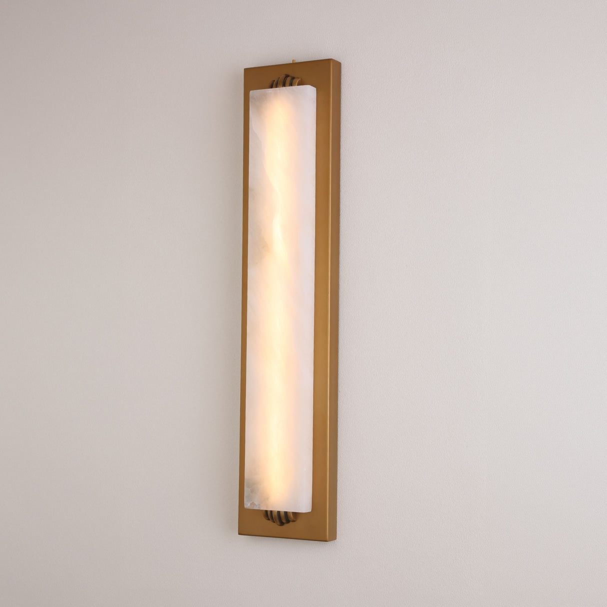 Valora vertical alabaster wall light illuminated on a pale wall, showing natural stone veining within a slim brushed brass rectangular frame.
