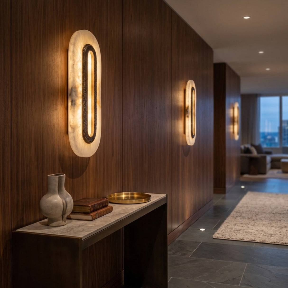 Ovaline LED Oval Alabaster Wall Light 46cm in Brushed Brass, glowing stadium-shaped alabaster plates with hammered brass rings lighting a wood-panelled hallway.
