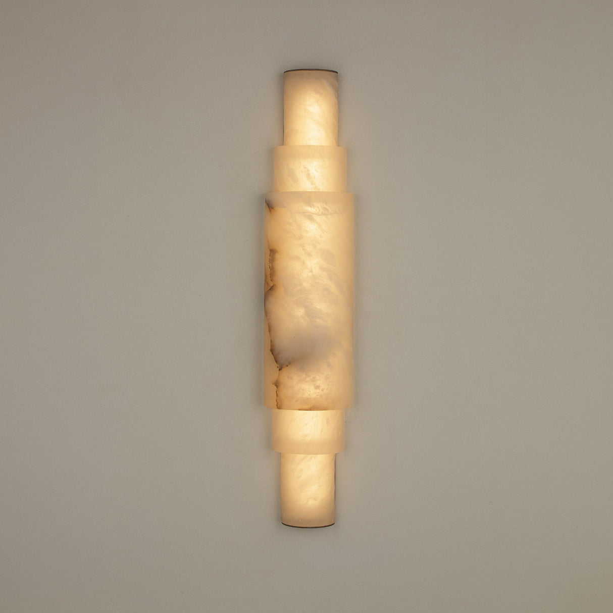 Niori Aurelia LED wall light in natural Spanish alabaster with stacked cylindrical sections and brushed brass end caps, illuminated against a neutral wall.