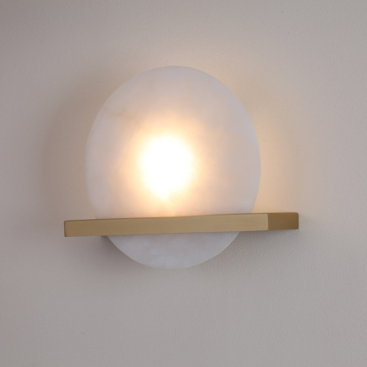 Niori Caldris circular alabaster wall light illuminated with warm LED glow, bisected by a brushed brass bar, mounted on a neutral wall.