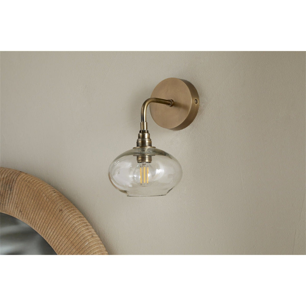 Otoro Recycled Glass Wall Light in Clear, with a round brass wall plate, curved brass arm, and flattened clear glass shade exposing the bulb, shown on a neutral wall above a rattan mirror edge.