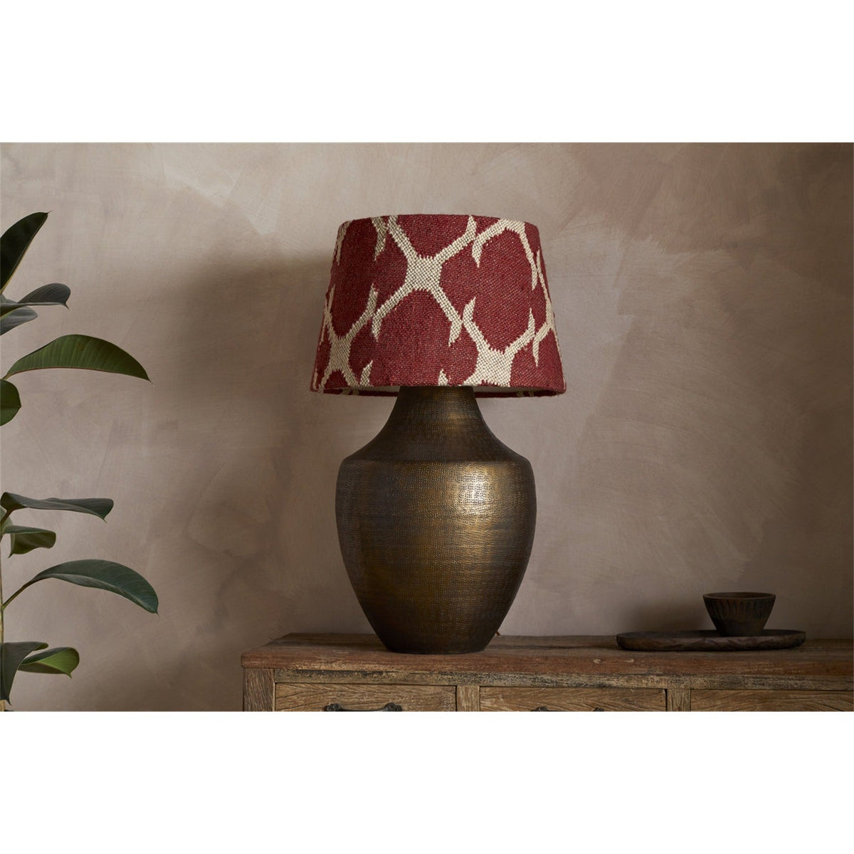 Haadu Large Jute Kilim Lampshade in rust and natural wool, shown atop a hammered bronze-finish lamp base on a wooden sideboard.