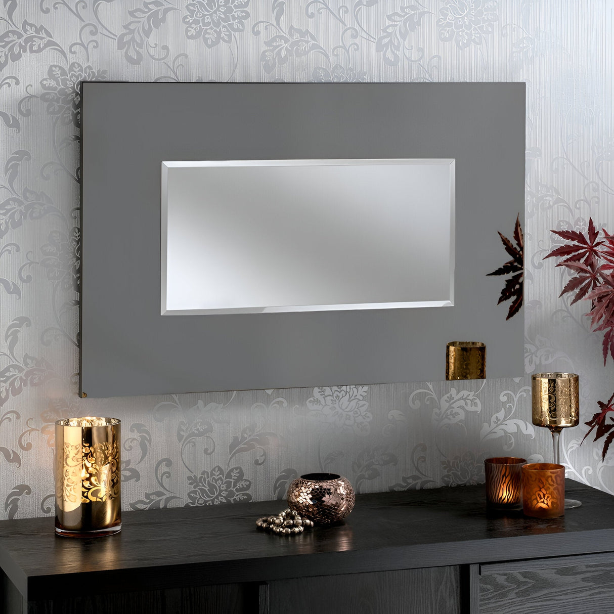 A contemporary Clarius Rectangle Wall Mirror in grey with dimensions 92x61cm hangs on a floral-patterned wallpapered wall. Below, a table holds decorative items including a gold candle holder, a small beaded ornament, and a dark vase with leafy branches.