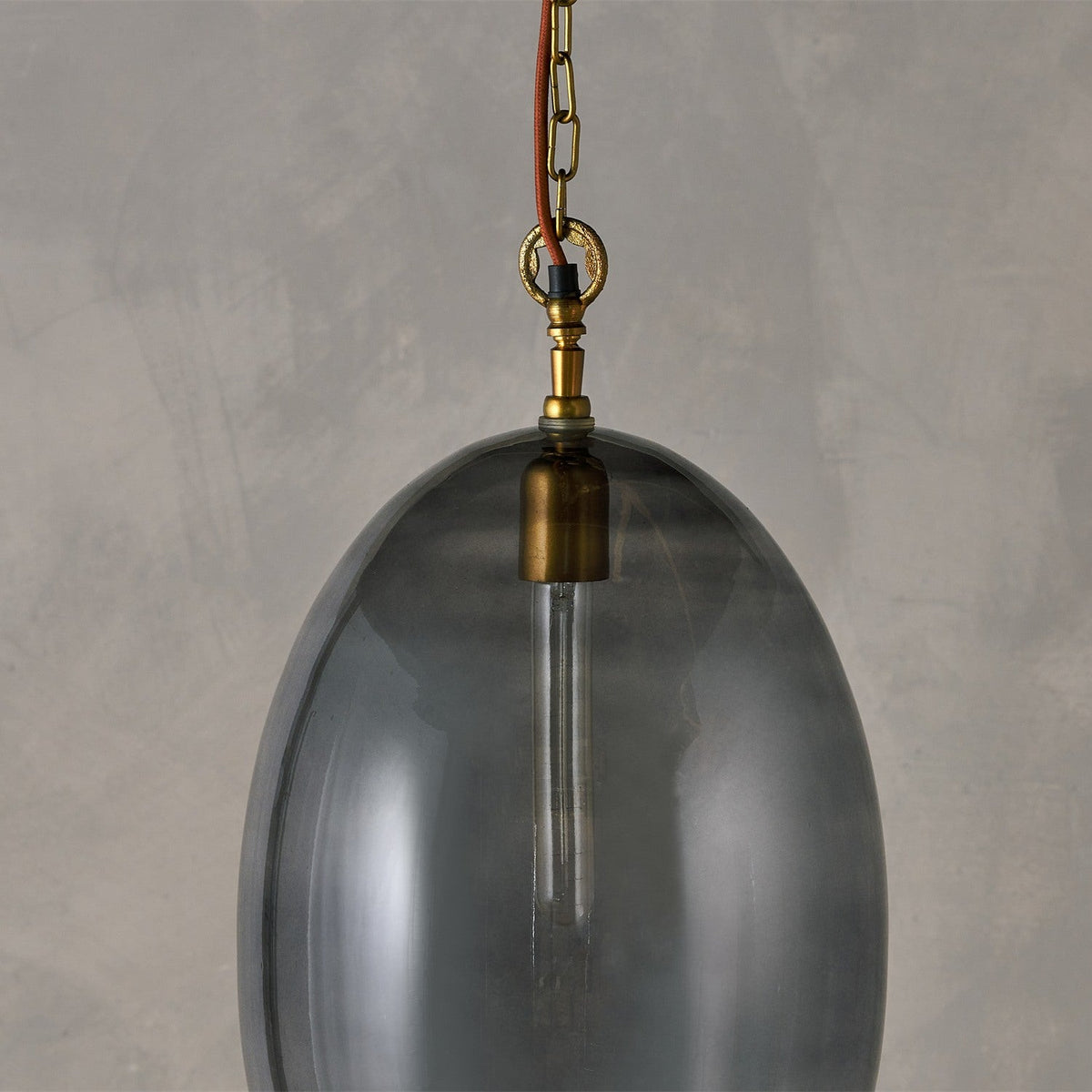 Otoro Large Oval Recycled Glass Pendant Light in smoke, close-up of the egg-shaped shade with brass collar, chain, and rust fabric flex against a plain grey background.