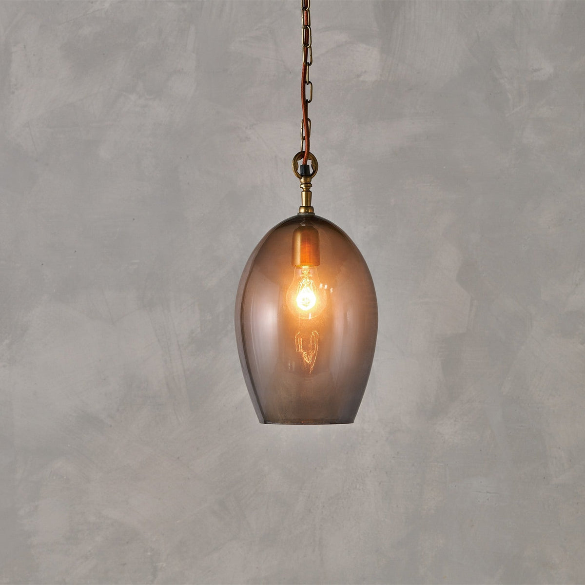 Otoro Large Oval Recycled Glass Pendant Light in smoke, an egg-shaped shade with brass collar and chain on rust fabric flex, lit bulb visible, set against a grey background.