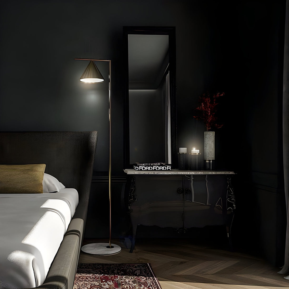 A dimly lit, elegantly designed bedroom features the Odin Floor Lamp - Brass beside the bed, a decorative rug, and a dark ornate dresser displaying a mirror, lamp, red flowers, candles, and books.