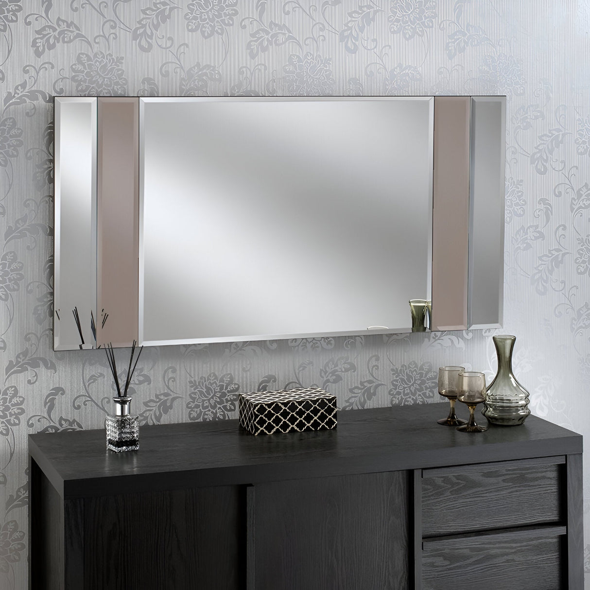 In a contemporary living room, elegant floral wallpaper sets the stage for the Brillia Rectangle Wall Mirror Bronze - 116x61cm, which boasts striking geometric accents. Positioned above a sleek black wooden sideboard, it harmonizes beautifully with a glass vase filled with reeds, a decorative box, and two intricately designed glasses, achieving coordinated modern décor.