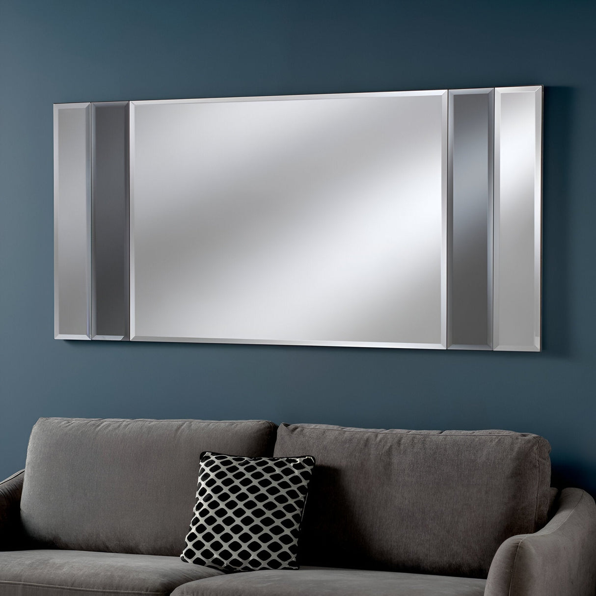 A Brillia Rectangle Wall Mirror in Grey, measuring 116x61cm, is mounted on a dark teal wall above a gray couch, elevating the modern home decor. A decorative cushion with a black and white pattern introduces contrast to the minimalist style, while the mirror's sleek design seamlessly blends into the setting.