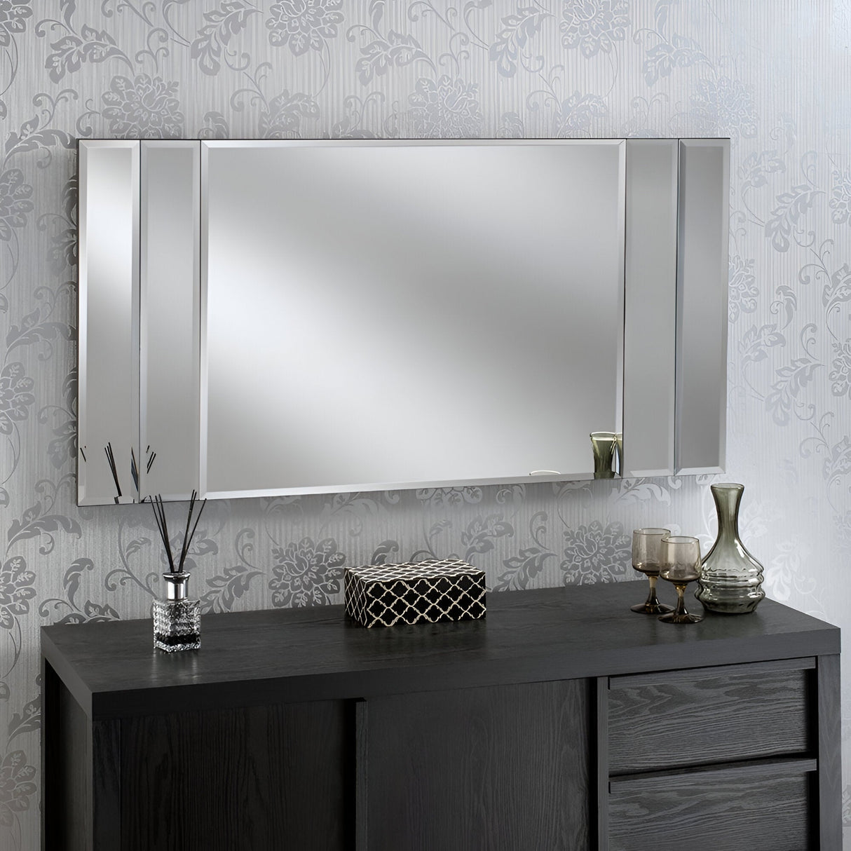 A Brillia Rectangle Wall Mirror in silver, measuring 116x61cm, adorns a patterned wallpapered wall. Beneath it, a dark wood console table exudes modern luxury, displaying decorative items such as a reed diffuser, a small patterned box, and three glass candle holders.