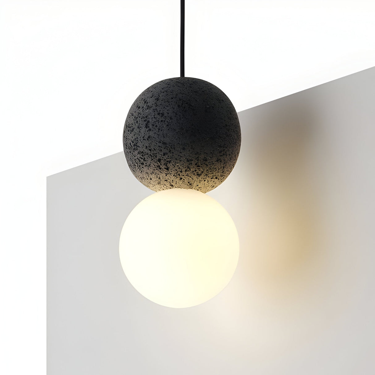 The Origo Concrete Pendant Light - Black & White features a volcanic rock-textured black sphere atop a glowing opal glass diffuser, suspended over a minimal white surface with soft shadows and light.