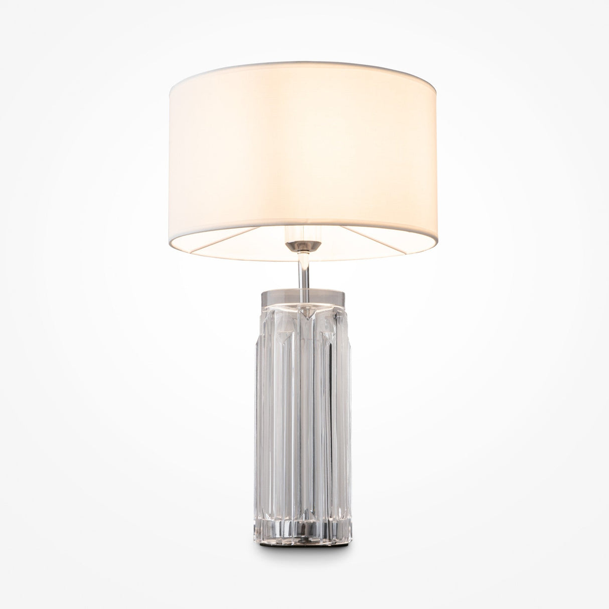 The Muse Table Lamp - Chrome With White Shade features a cylindrical, transparent glass base with vertical ridges, ideal for modern interiors. A wide, cream-colored fabric shade emits a warm glow and is elegantly set against a plain white background.