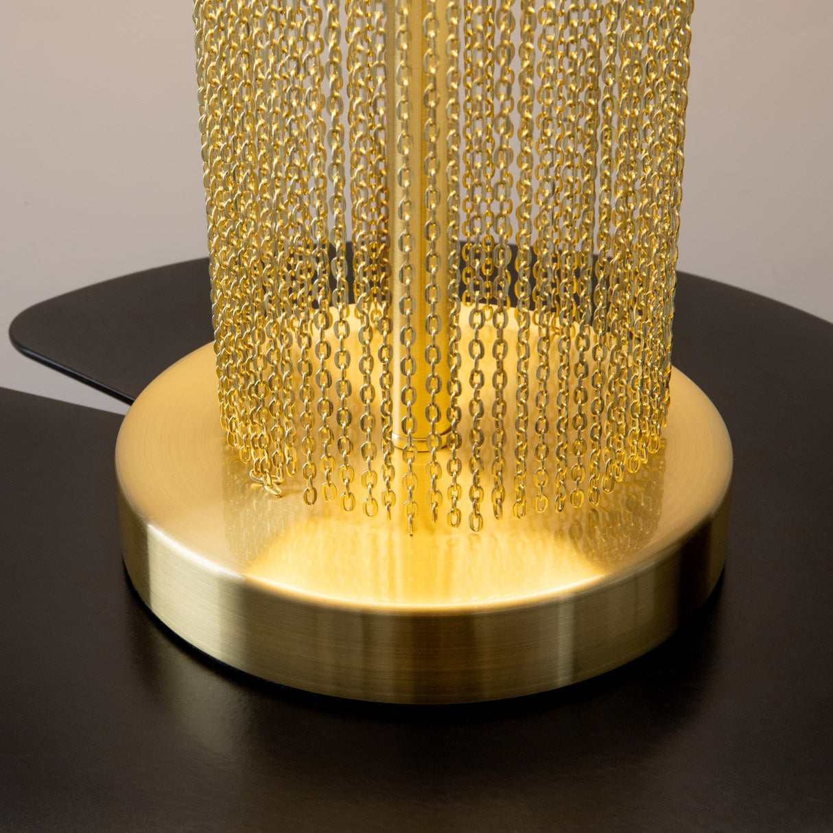 Close-up of the Impressive Table Lamp, showcasing its luxurious gold cylindrical base and cascading chains creating a curtain effect. The gold accents reflect soft light on the black surface, with a complementing white shade adding elegance.