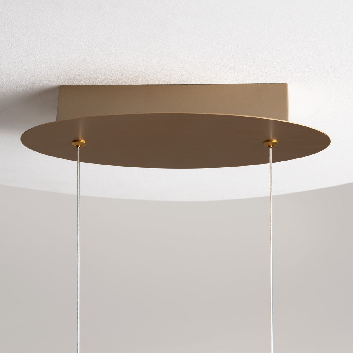 The Curve LED Loop Suspension Light - Gold features a modern design with a flat, circular beige shade and gold finish. It hangs from a rectangular base on two thin wires with an integrated LED light source, elegantly set against a white ceiling.