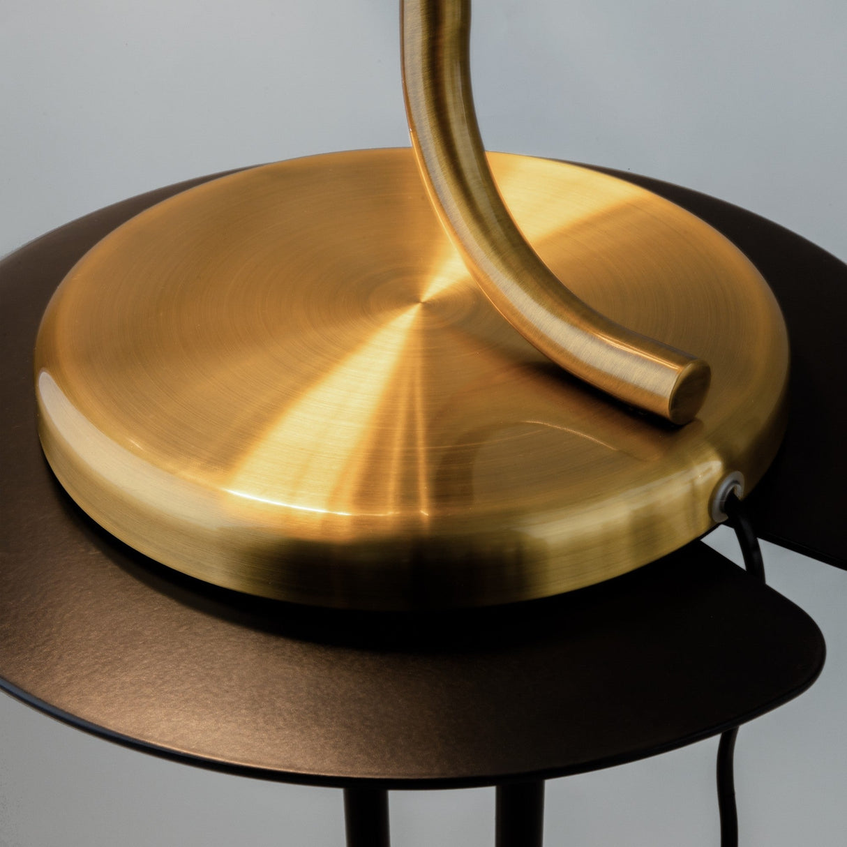 Close-up of the Cabaret LED Table Lamp - Brass by Pierre Gonalons, featuring a modern brushed gold lamp base with a sleek, curved stem. Positioned on a black surface, its energy-efficient lighting is complemented by a side power cord.