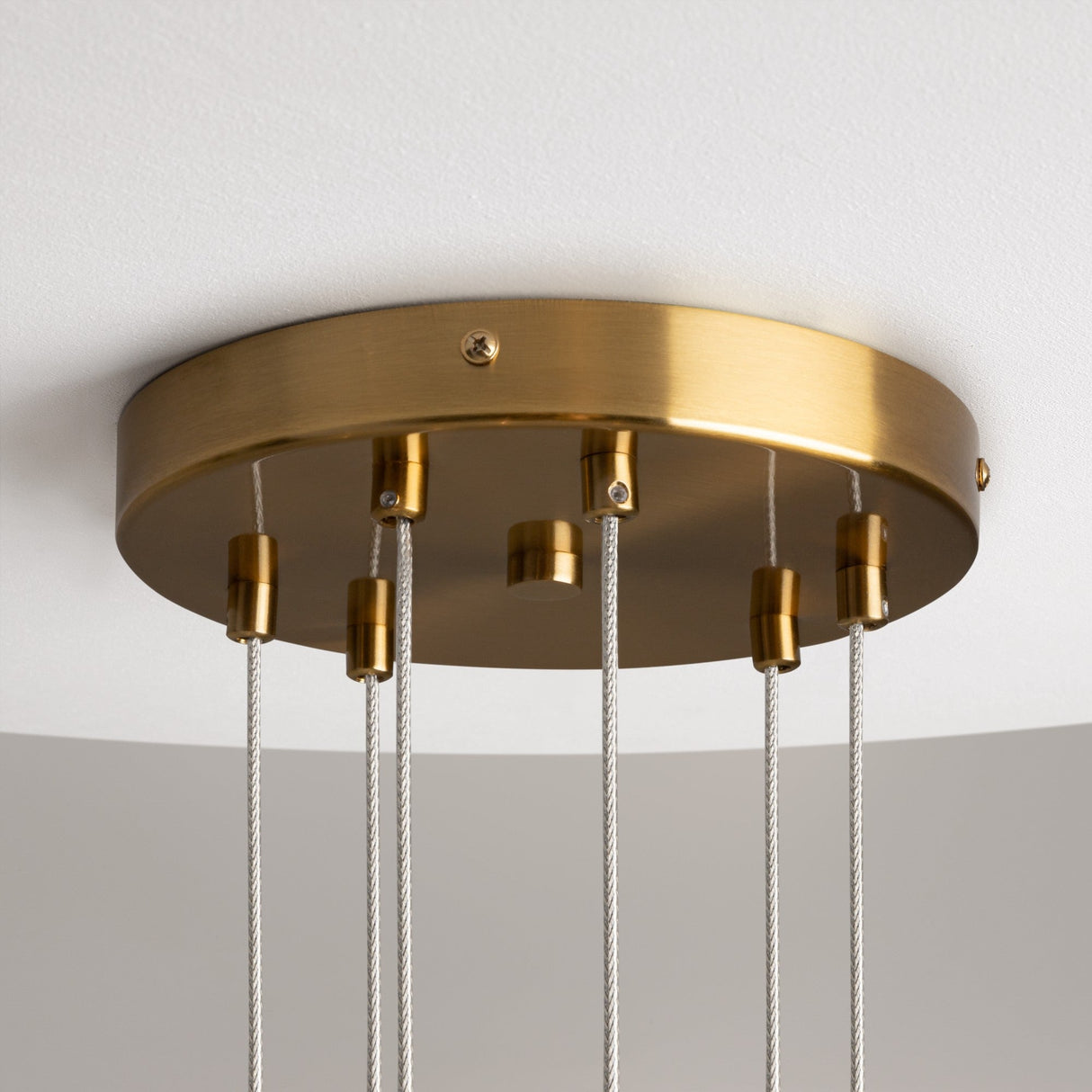 Detailed view of the Mood 6 Light Suspension, featuring a gold circular base in brass. Thin cables descend to support the clear glass shades (not shown), with adjustable height and ceiling-mounted on white backdrop.