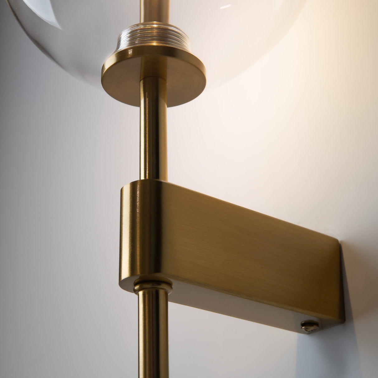 Close-up of the Mood Wall Light - Brass With Clear Round Glass. This elegant fixture showcases a metal arm extending from a rectangular wall mount, supporting a round glass shade, creating a warm ambiance.