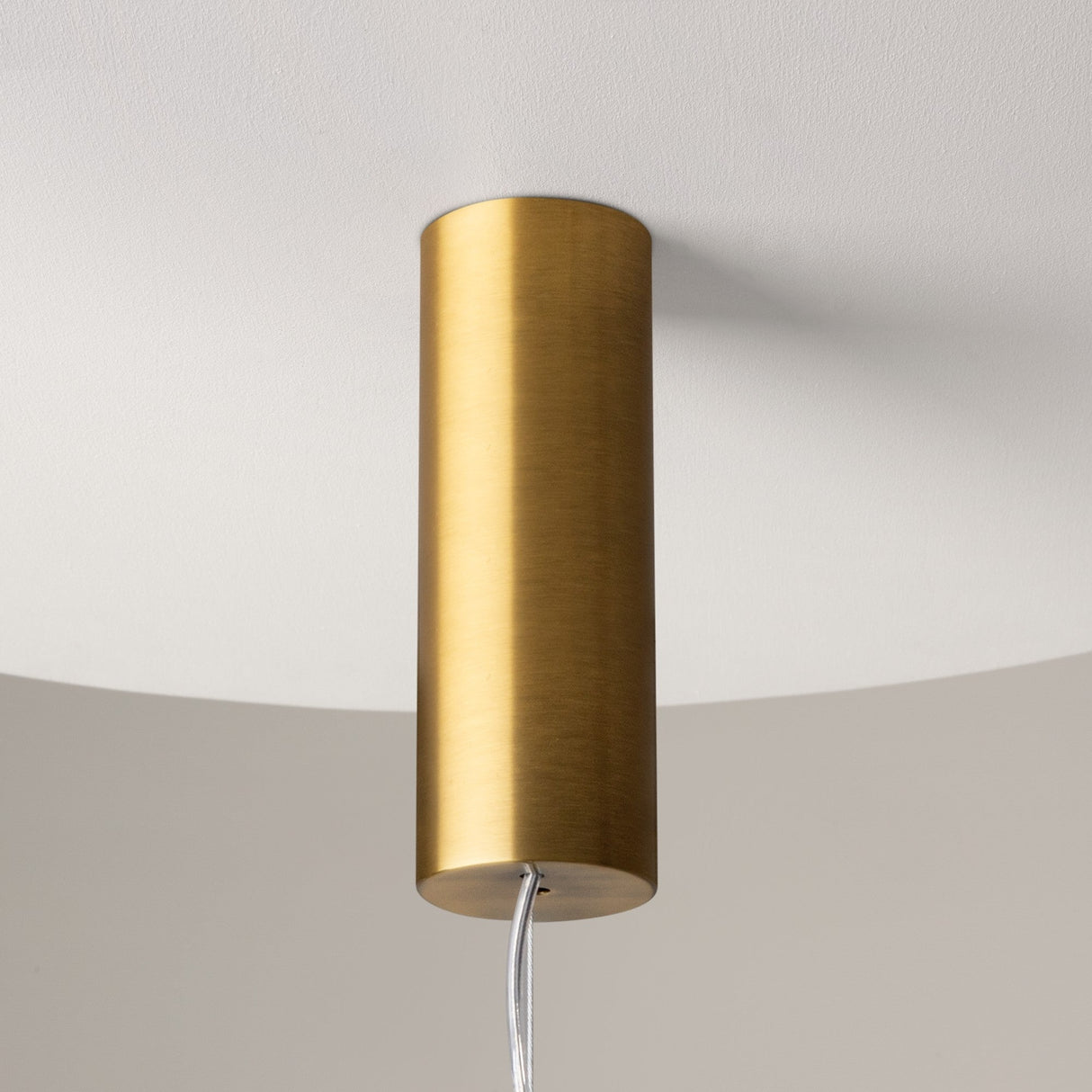 A close-up of the Cosmo LED Linear Suspension Light in brass with clear glass, featuring a modern cylindrical design and celestial elements, hangs elegantly from a white ceiling with its minimalist brushed metal finish and visible cable.