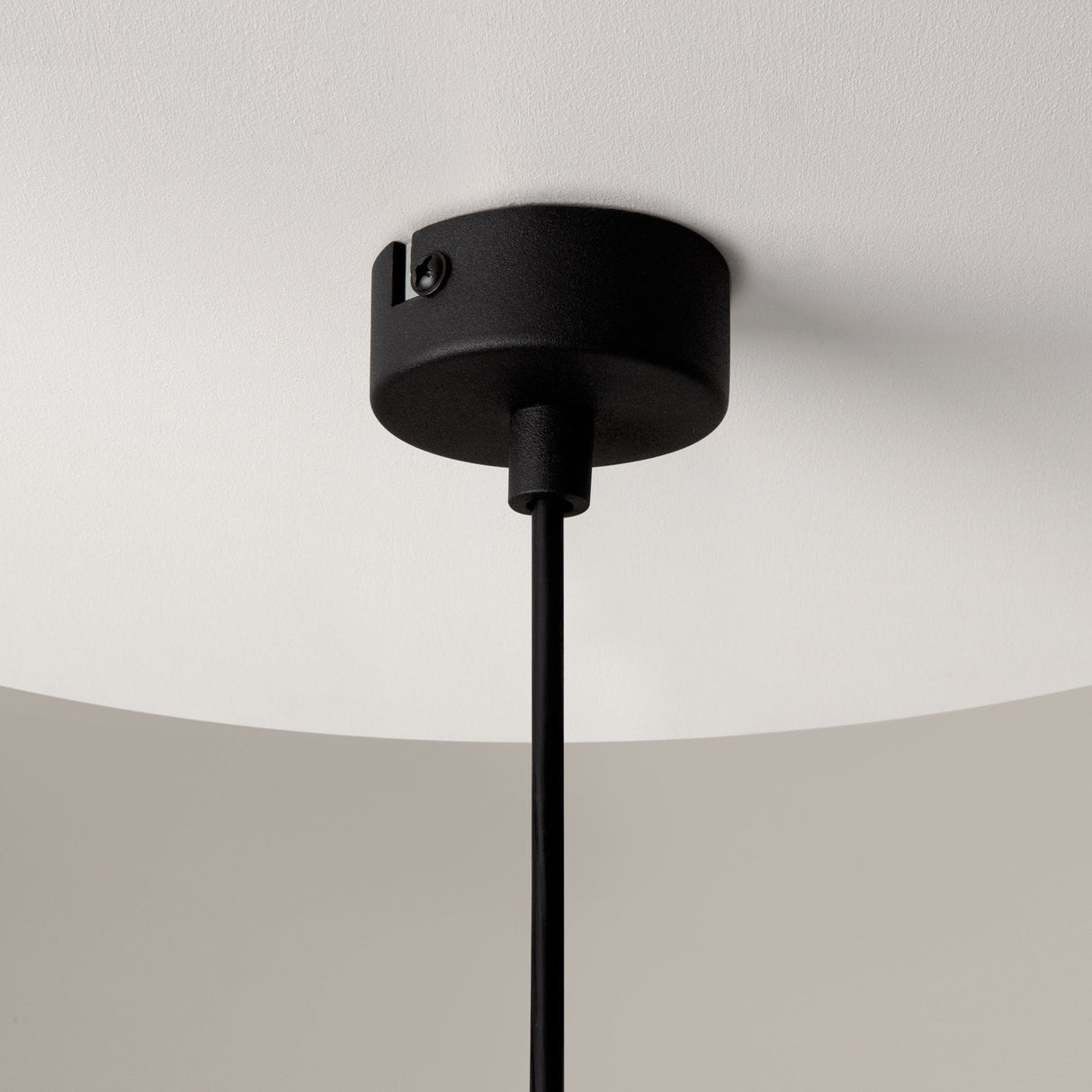 The image features a Ros 1 Light Pendant with a black matte base on a white ceiling. A black cord dangles an elegant amber glass shade, showcasing its minimalistic and modern design.