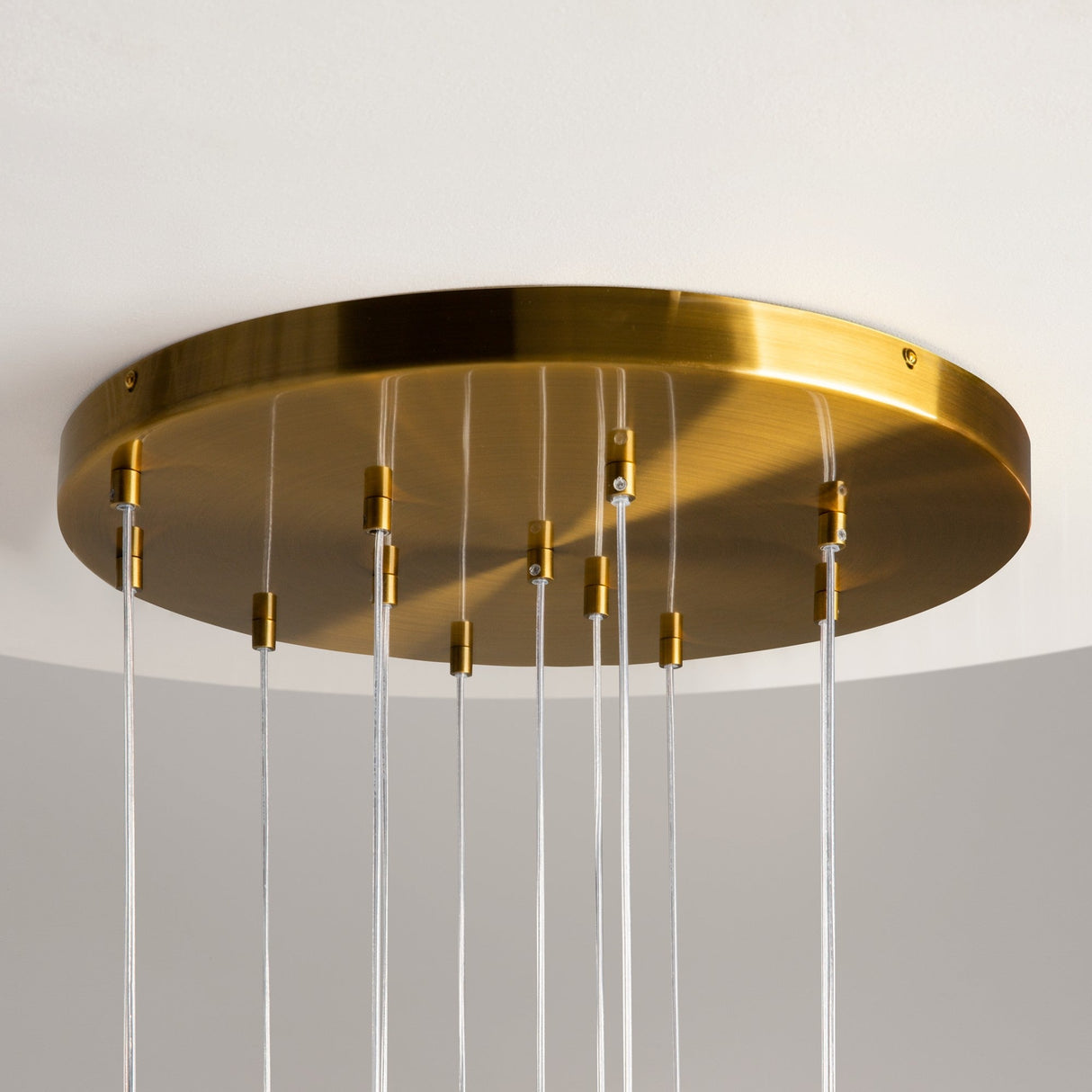 The Frozen 12 Light Spiral Pendant features a round brushed brass plate, solid glass lampshades, and transparent wires that elegantly support small brass elements in a minimalist design using metallic and clear materials.