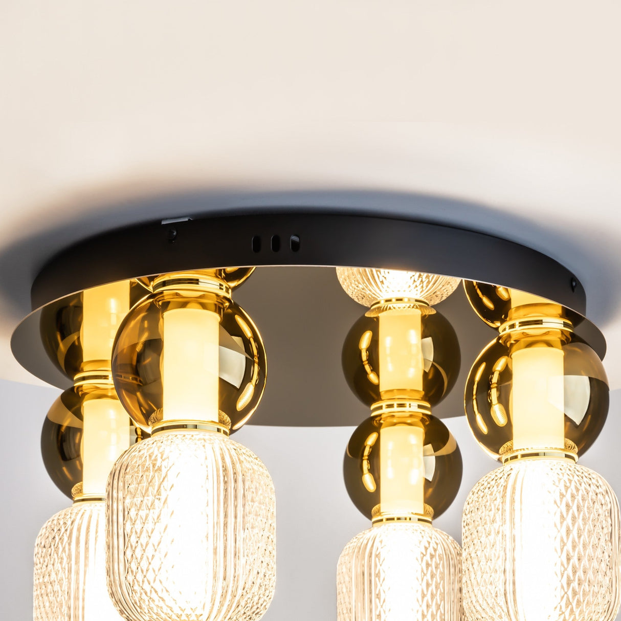 Close-up of the Drop LED Flush Light 3000K - Gold, an elegant fixture with polished black metal and gold accents. It has four cylindrical textured glass lampshades, emitting warm energy-efficient light, hanging from spherical golden connectors against a white ceiling.