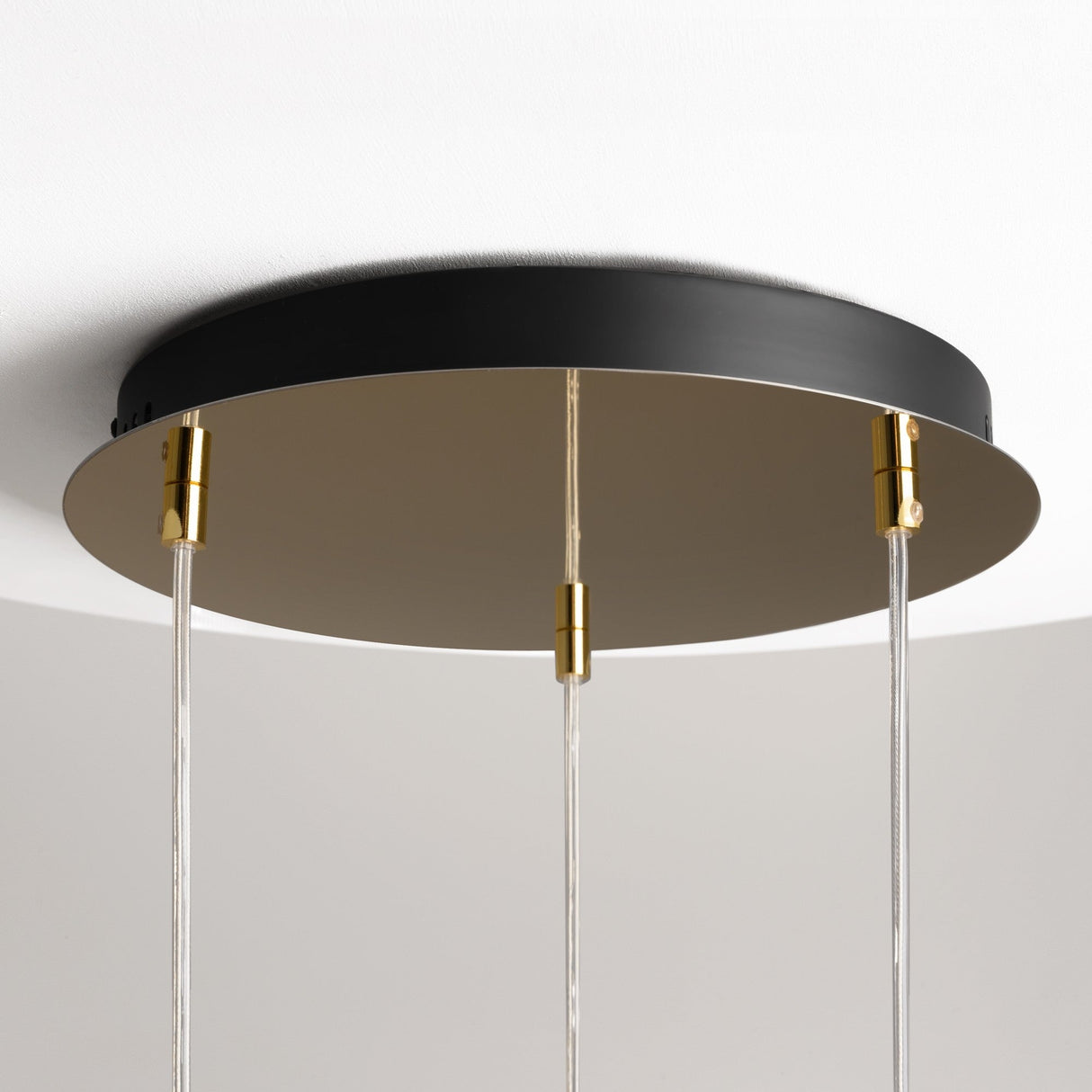 A close-up of the Drop LED Cluster Pendant Light 33W 3000K - Gold reveals a modern fixture with a round, black and gold base. It features three clear rods hanging vertically, each with refined gold fittings, providing energy-efficient lighting against a plain, light-colored ceiling.