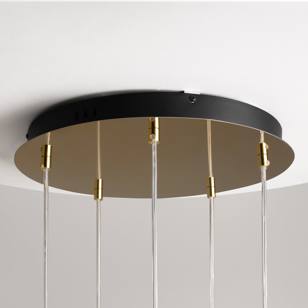 A close-up showcases the Drop LED Cluster Pendant Light 80W 3000K - Gold, featuring transparent rods and a round black base. Gold connectors affix to a sleek metal frame, creating an elegant design. This energy-efficient fixture beautifully complements the white ceiling.