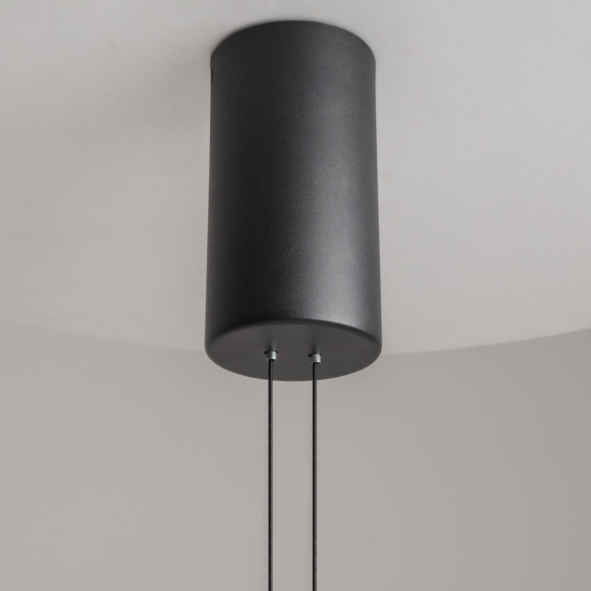 Close-up of the Ambience LED Round Flat Large Pendant Light in black, featuring a modern cylindrical design in Japandi style with two black cords against a plain, light-colored ceiling.