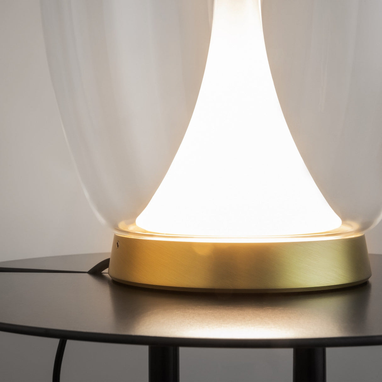 A close-up of the Splash LED Table Lamp 3000K 9W features a glowing white light under a sleek, transparent shade. The metallic gold base with clear glass accents contrasts against the black table, emitting a soft, warm glow for an elegant and minimalist ambiance.
