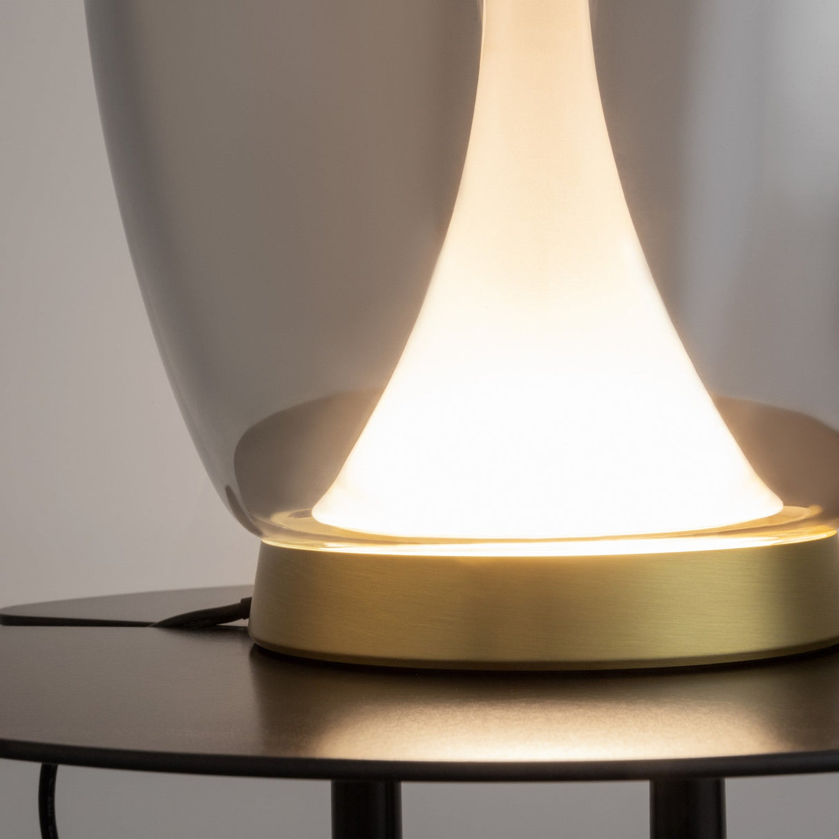 The Splash LED Table Lamp 3000K 9W in gold, with a frosted glass shade, emits a soft, warm glow. It features smoked ombre glass and contrasts elegantly against its metallic base on a dark wooden surface and neutral background.